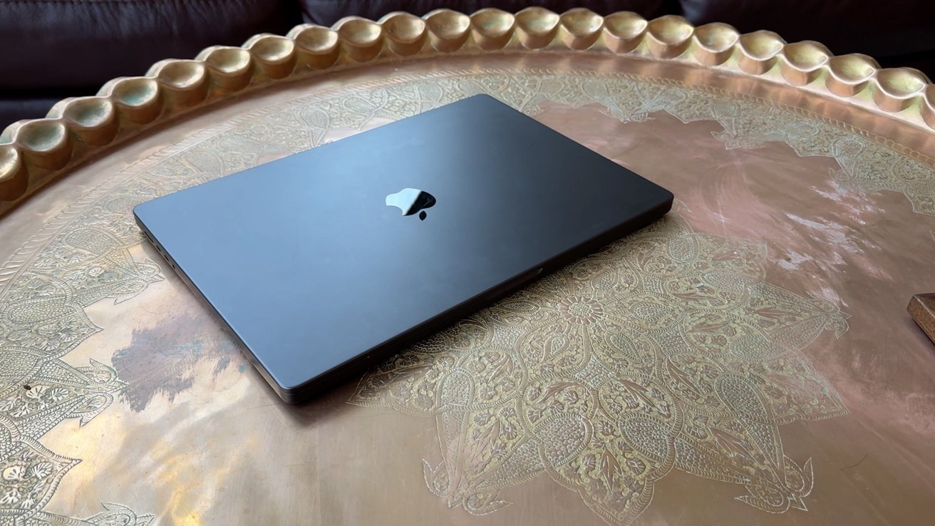 A closed black laptop on an ornate metallic gold-patterned table, with a brown sofa in the background.