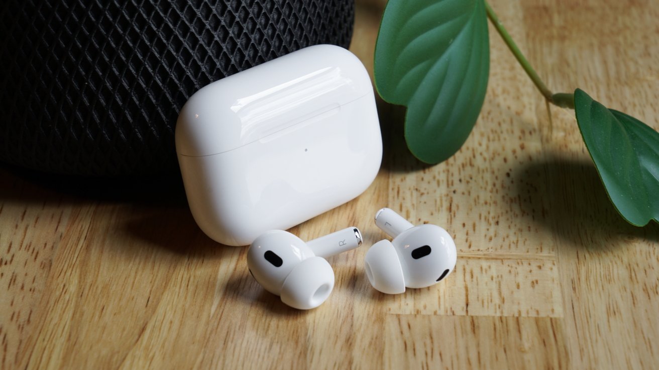 White wireless earbuds and charging case on a wooden surface, near a decorative green leaf and a black textured object in the background.