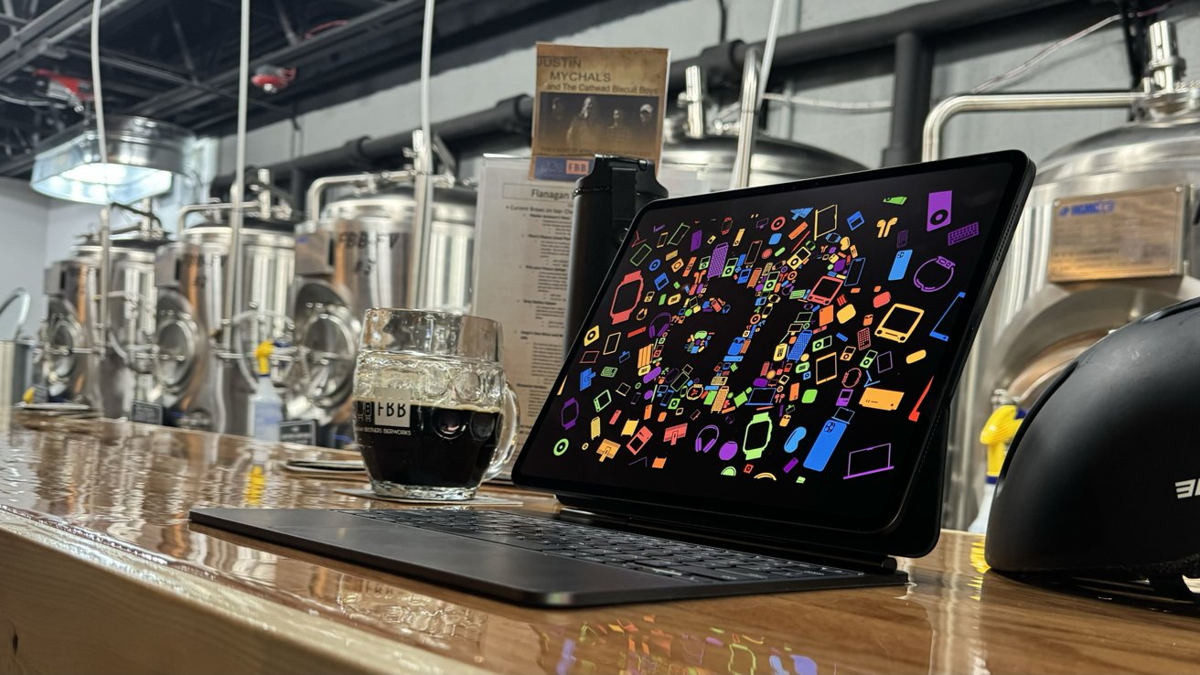 A tablet displaying colorful geometric shapes sits on a wooden table next to a filled beer glass, with a background of brewing tanks.