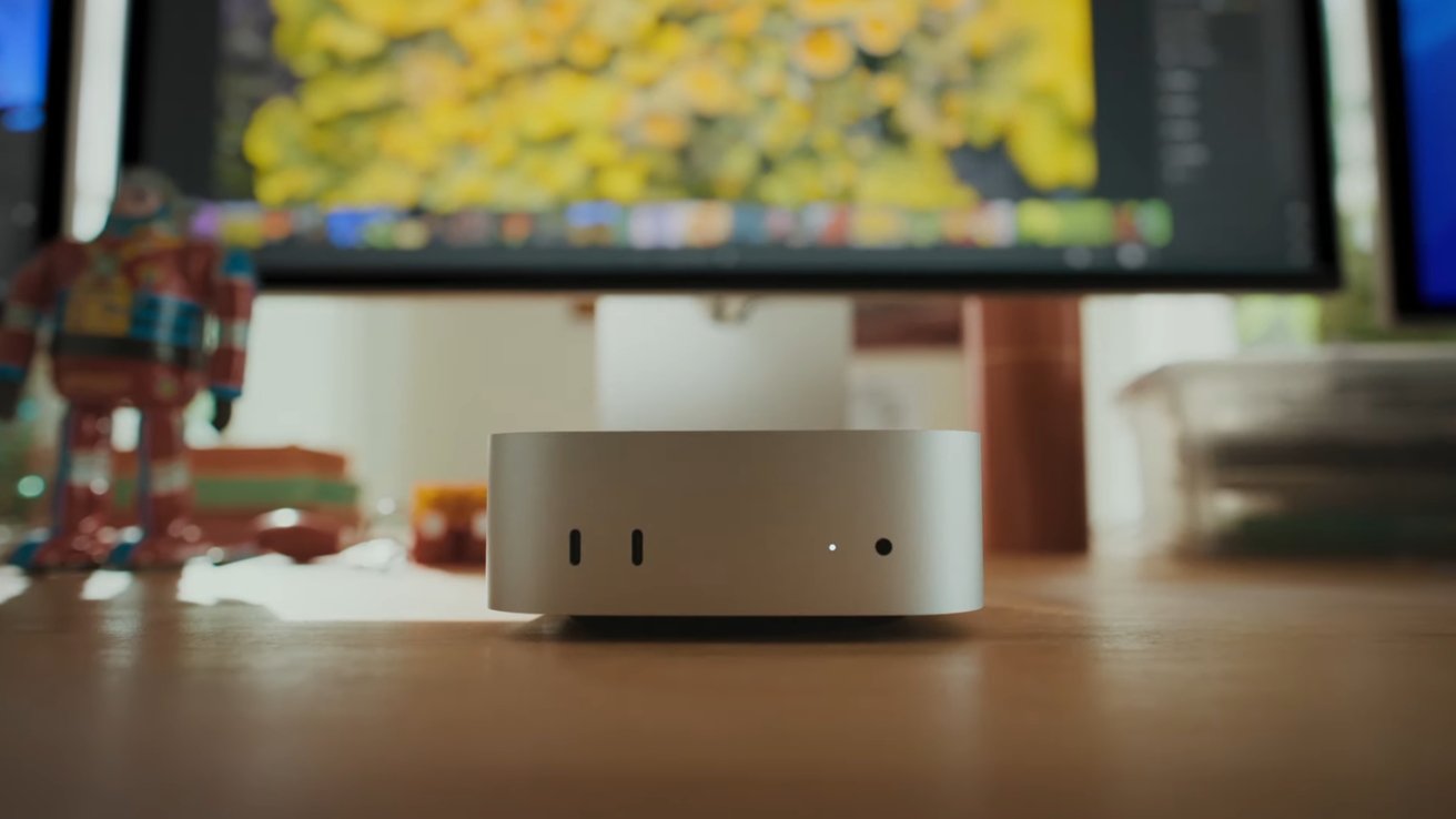 Small white computer device on a wooden desk, with a blurred monitor displaying yellow flowers in the background.