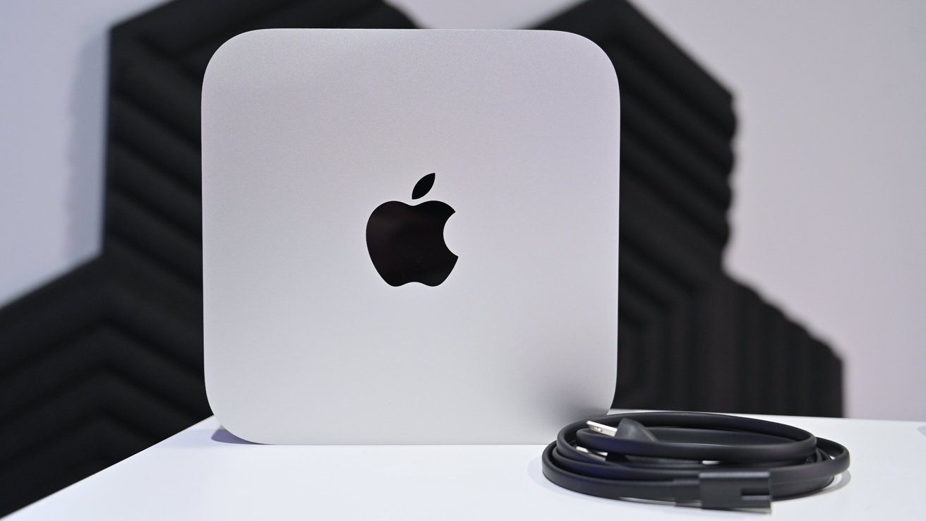 Silver square computer with an Apple logo, resting on a white surface next to a coiled black cable.