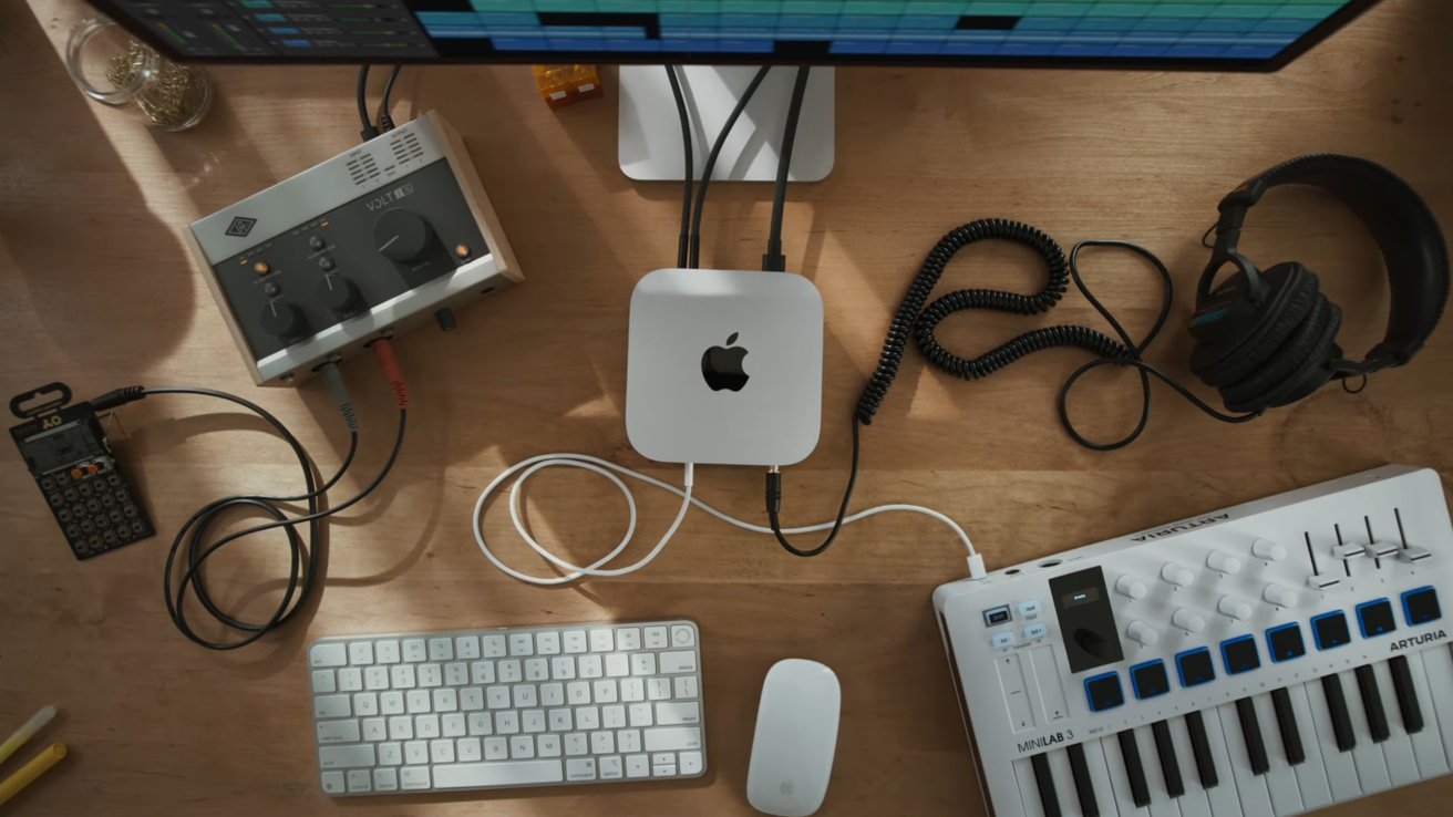 Music workstation with a keyboard, audio interface, headphones, and computer on a wooden desk. Cables connect various devices, and a monitor screen is partially visible.