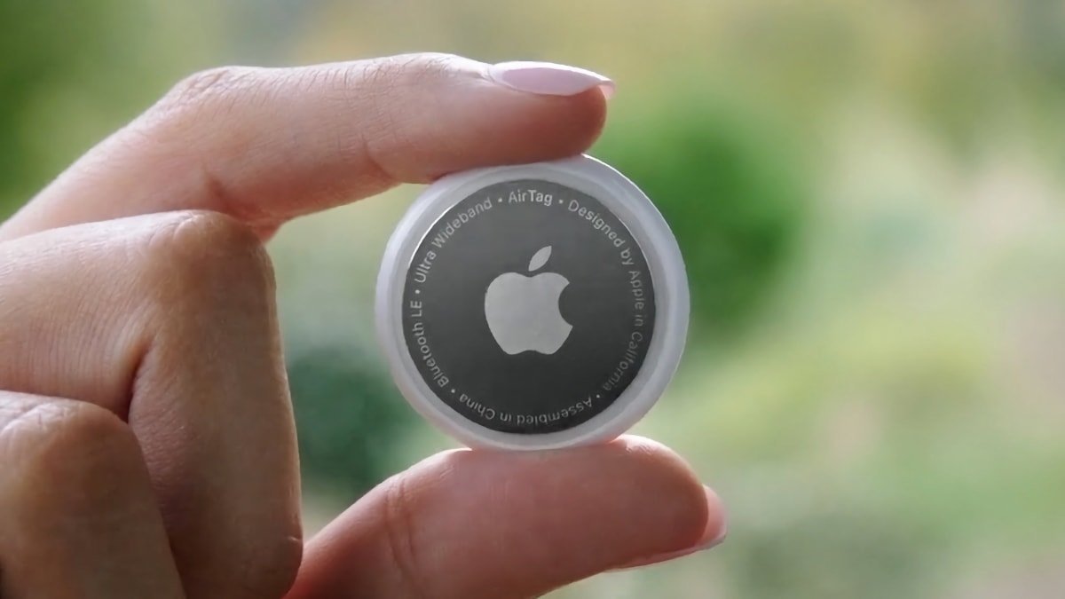 Hand holding a round Apple AirTag against a blurred greenery background, with the Apple logo and text.