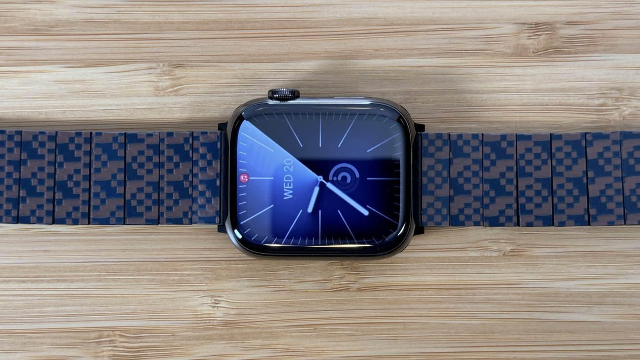 A smartwatch with a blue display showing time and date on a wooden surface with a pixel-patterned strap.