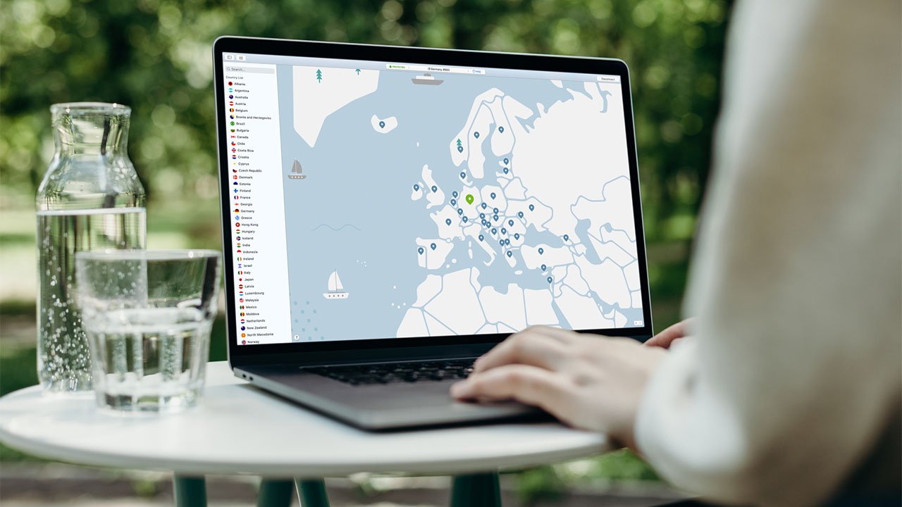 Person working on MacBook Pro displaying a map with location markers and available VPN servers on the screen, with water bottle and glass on outdoor table.