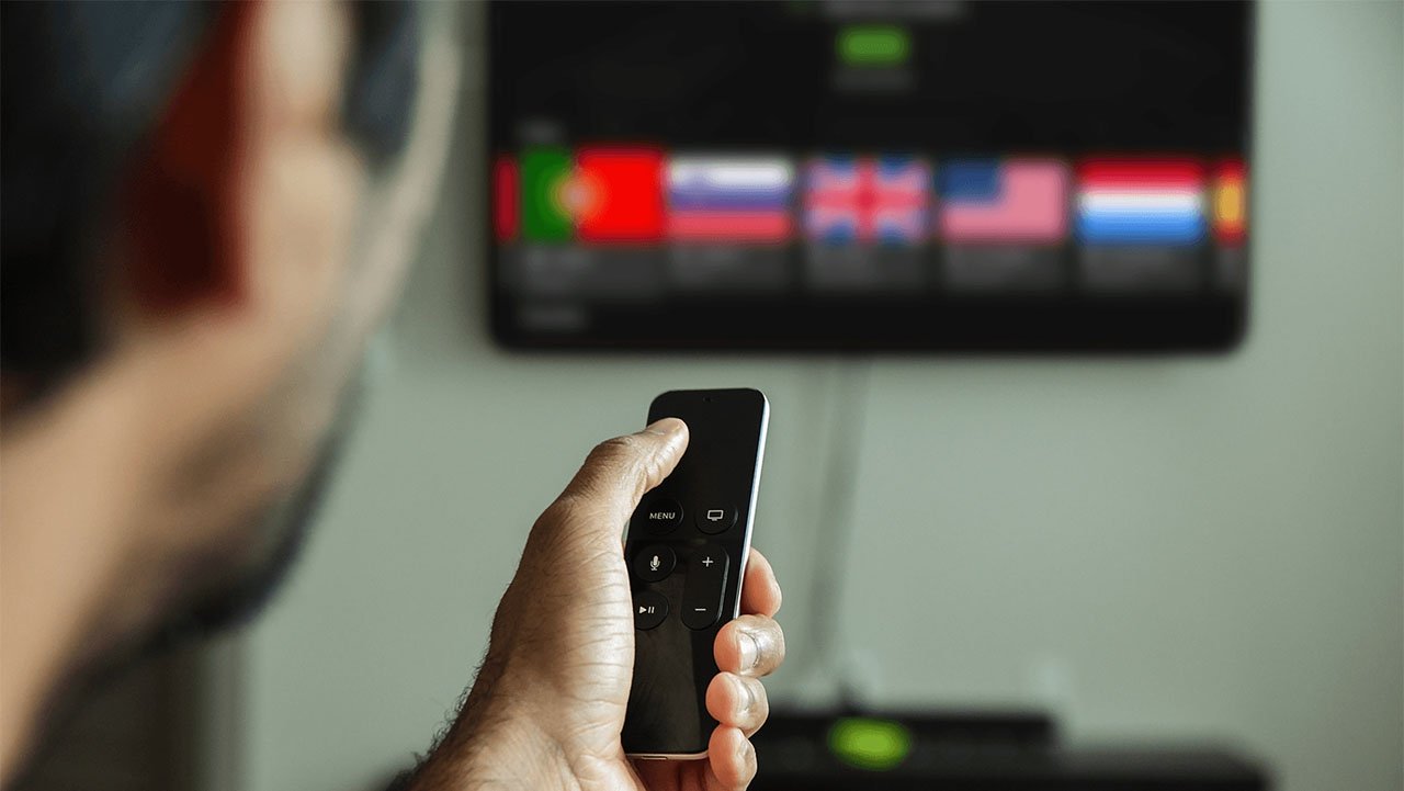 Person pointing a Siri Apple TV remote control at a television displaying a VPN selection of country flags in a row.