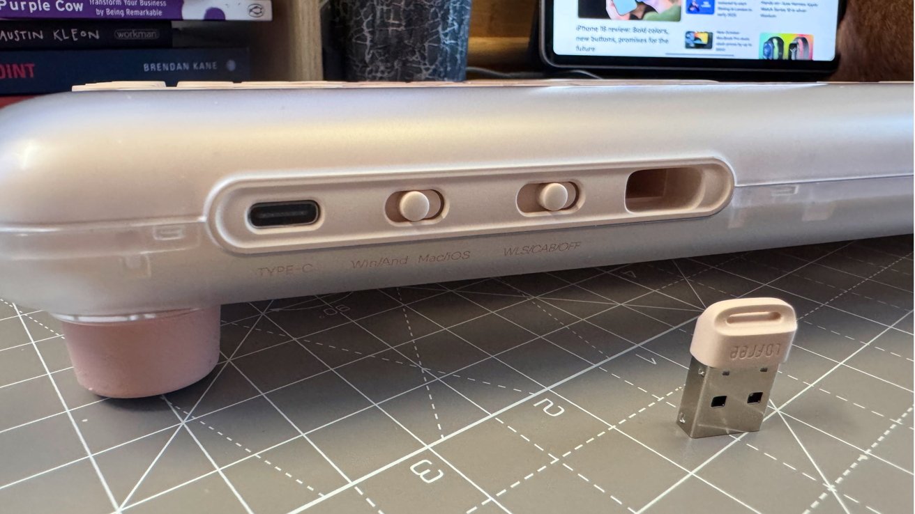 Close-up of a beige keyboard with USB-C port and switch options, next to a USB receiver on a grid-patterned desk mat. Books and a tablet are in the background.