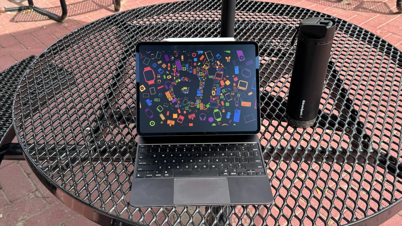 iPad Pro on a table outdoors in the Magic Keyboard. It is showing a colorful wallpaper filled with Apple products