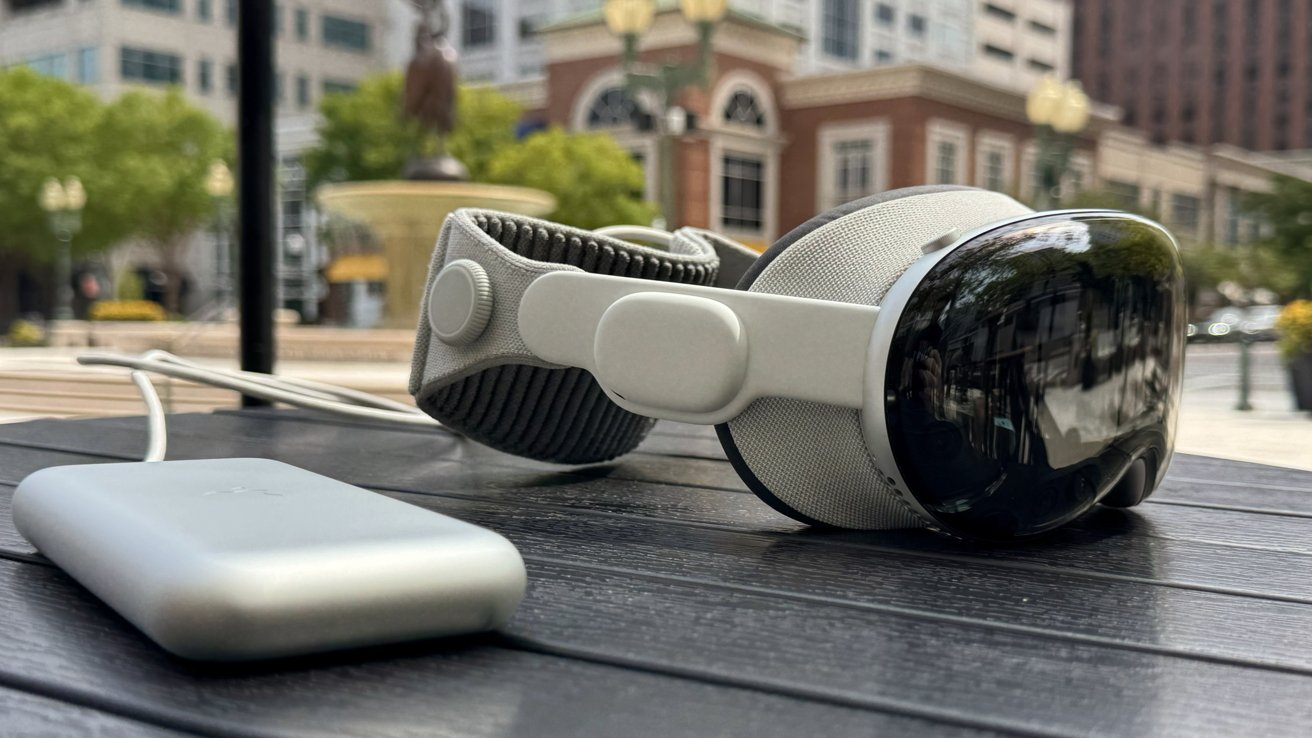 Apple Vision Pro sits on a table at a cafe with a fountain and buildings blurred in the background