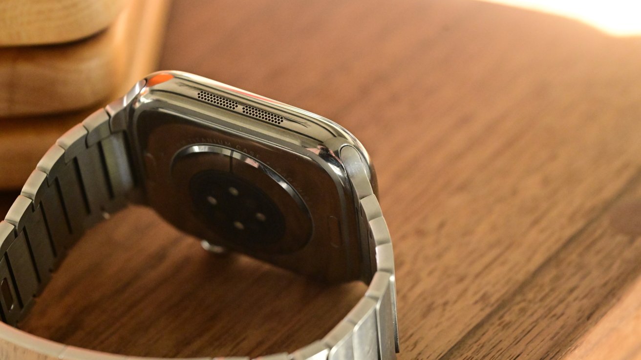 Smartwatch with a metallic band resting on a wooden surface, displaying the back sensor and speaker area.