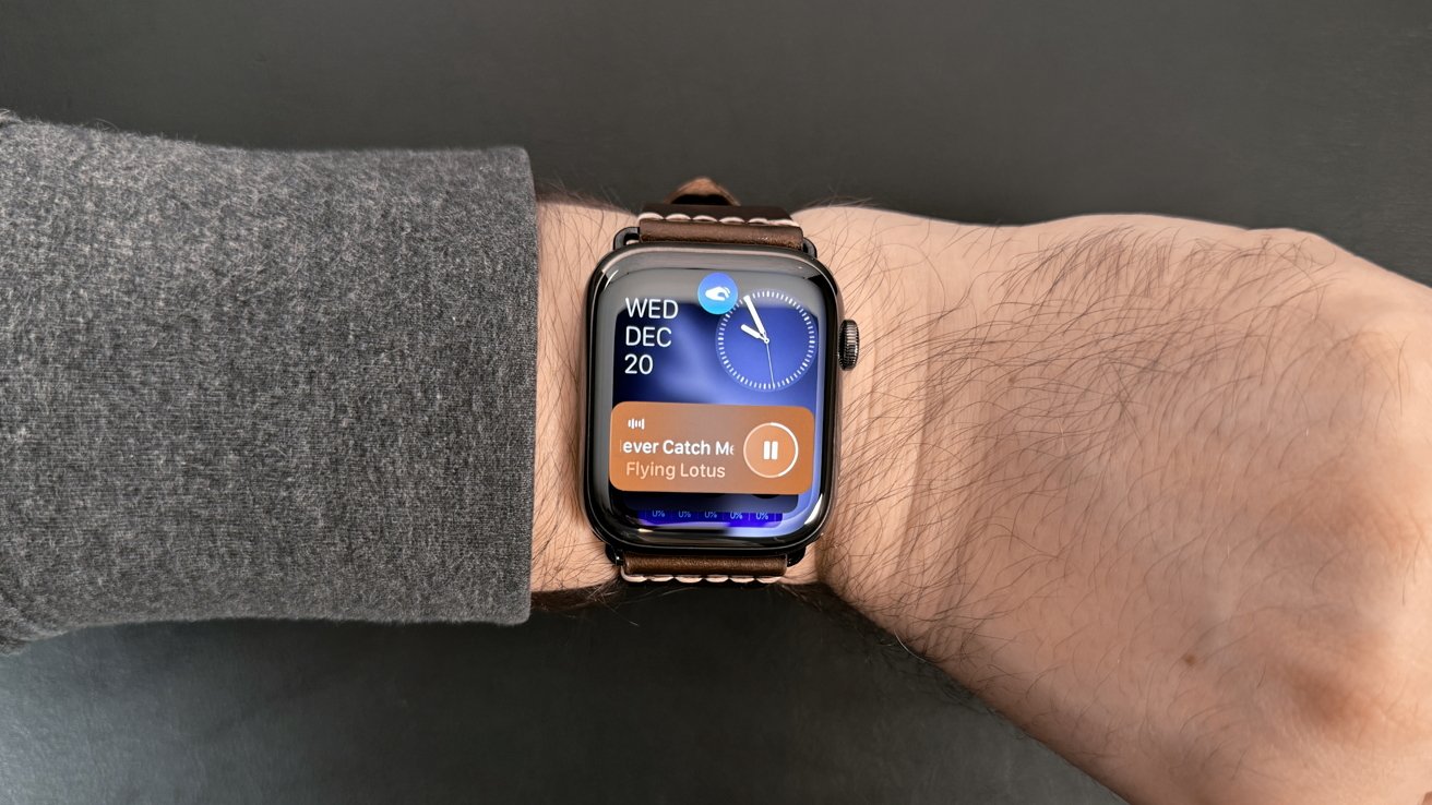 A smart watch on a wrist displays the date December 20, time, and a music player with the song 'Never Catch Me' by Flying Lotus.