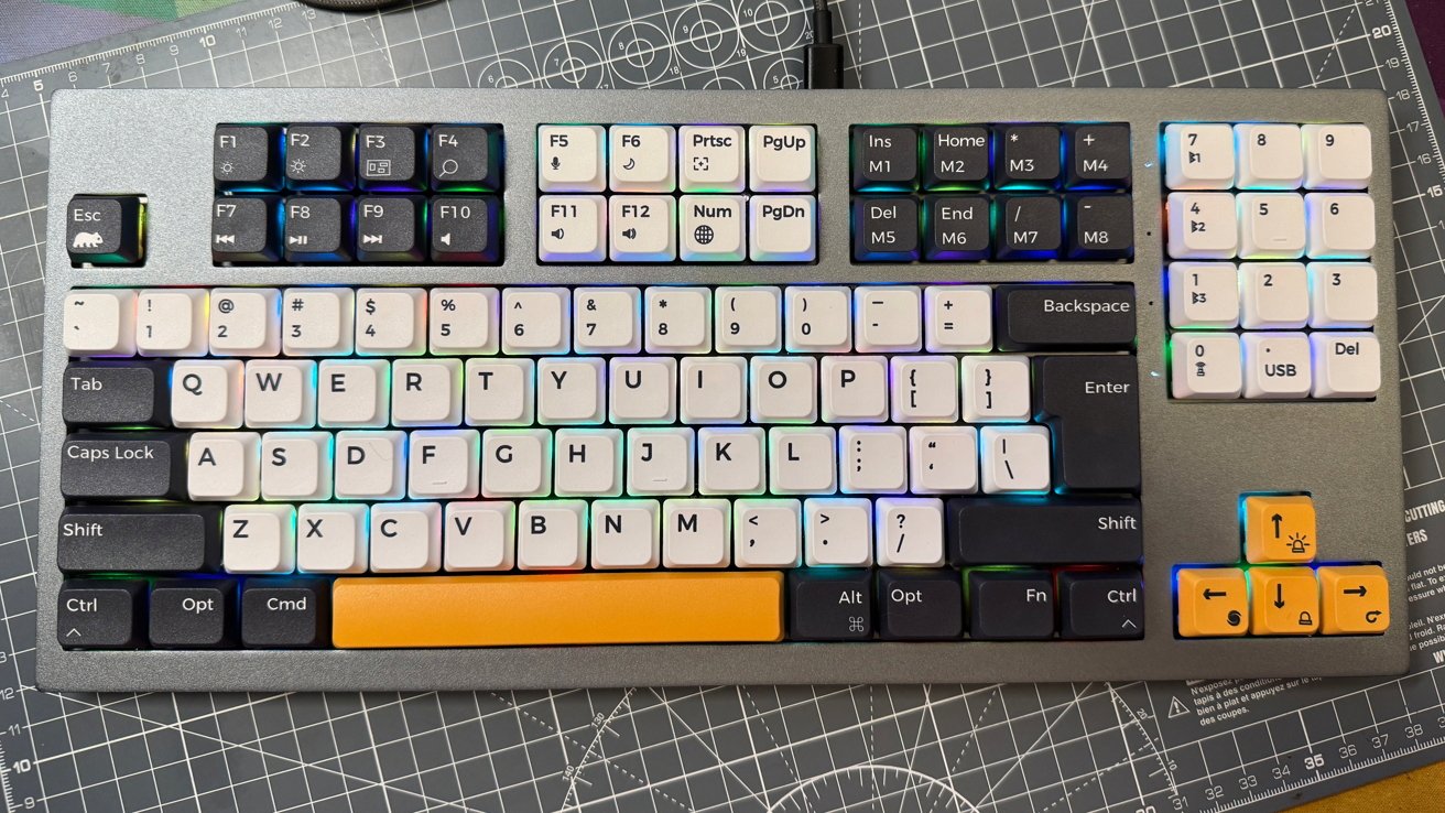 A mechanical keyboard with colorful backlit keys and a unique orange spacebar and arrow keys. It sits on a grid-patterned surface.