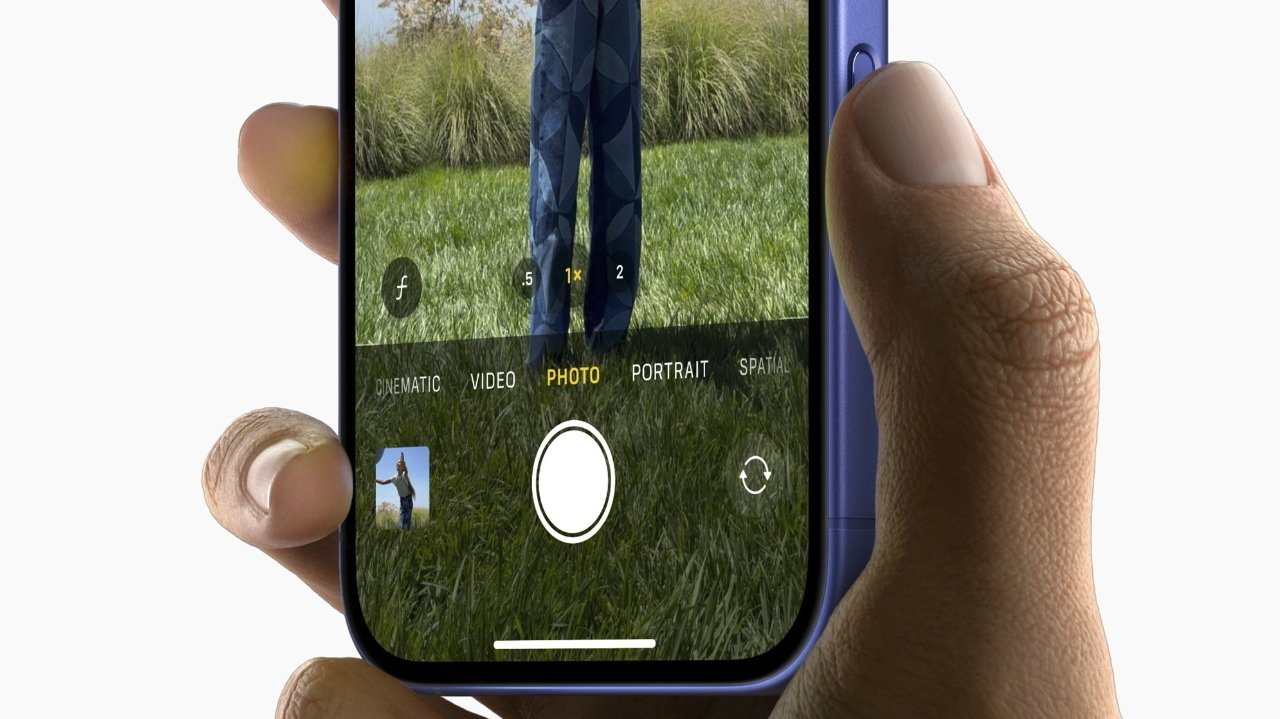 A hand holding a smartphone, displaying a camera app open in photo mode, capturing a person standing on grass.