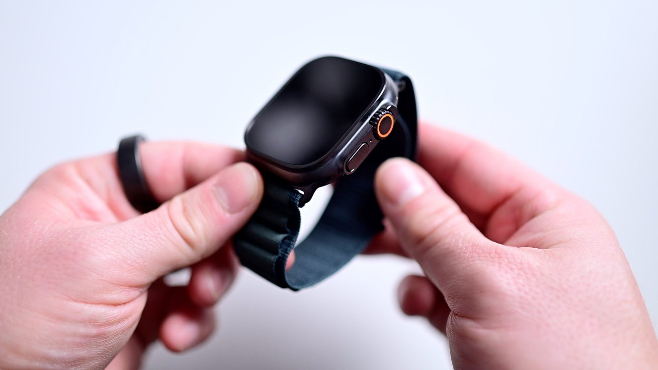 Hands holding a smartwatch with a dark screen and a blue strap, focusing on the side button and crown.