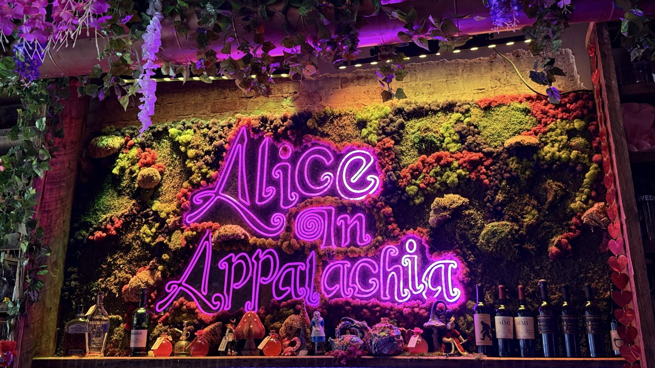 Neon sign reading 'Alice in Appalachia' against a green, mossy background, with hanging plants above and wine bottles below.