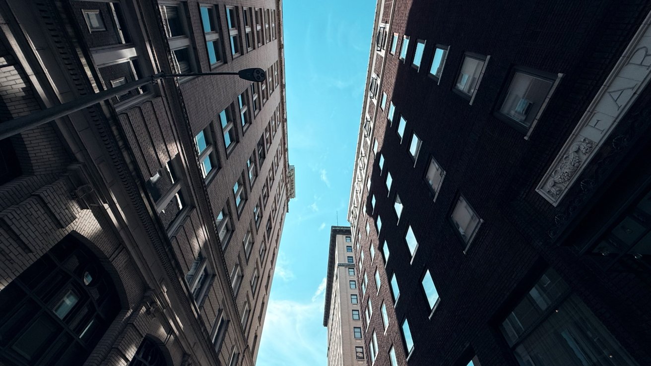 Tall buildings tower on both sides of a narrow street, looking up toward a bright blue sky with wispy clouds.