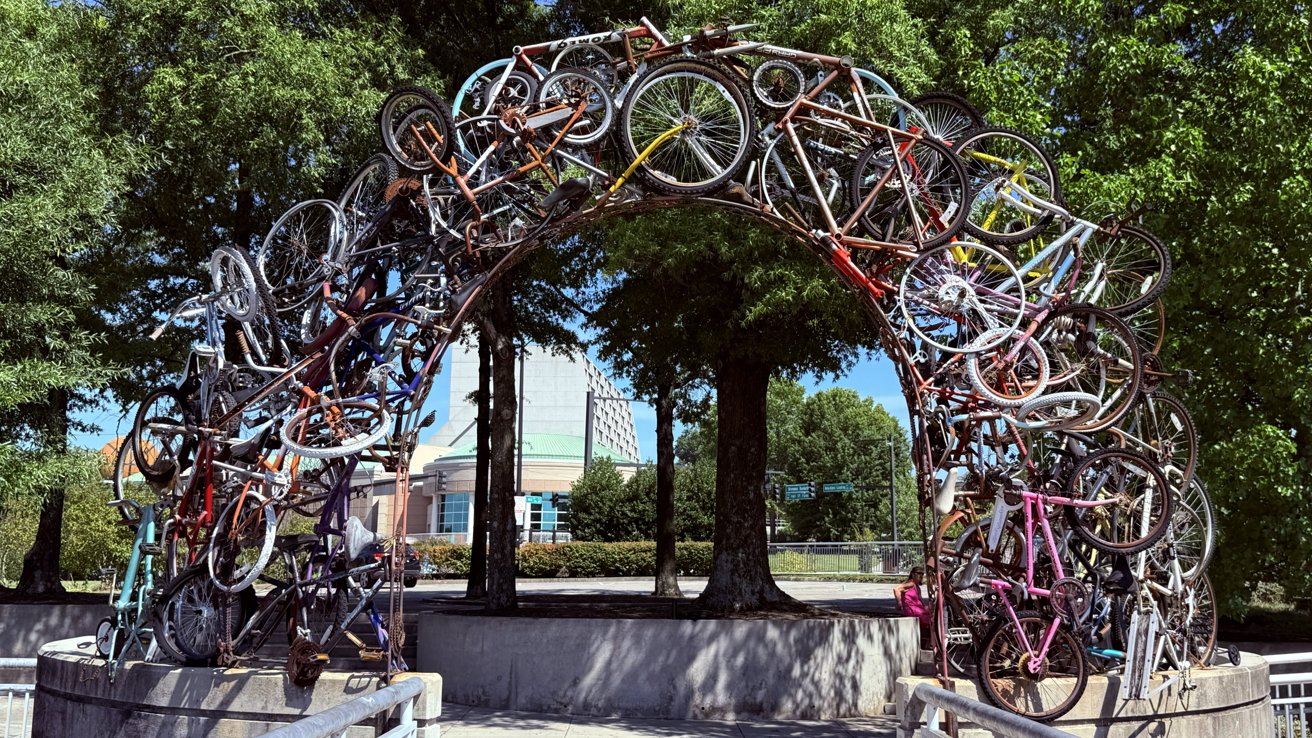 Arch sculpture made from numerous bicycles intertwined and stacked, set against trees and a building in the background.