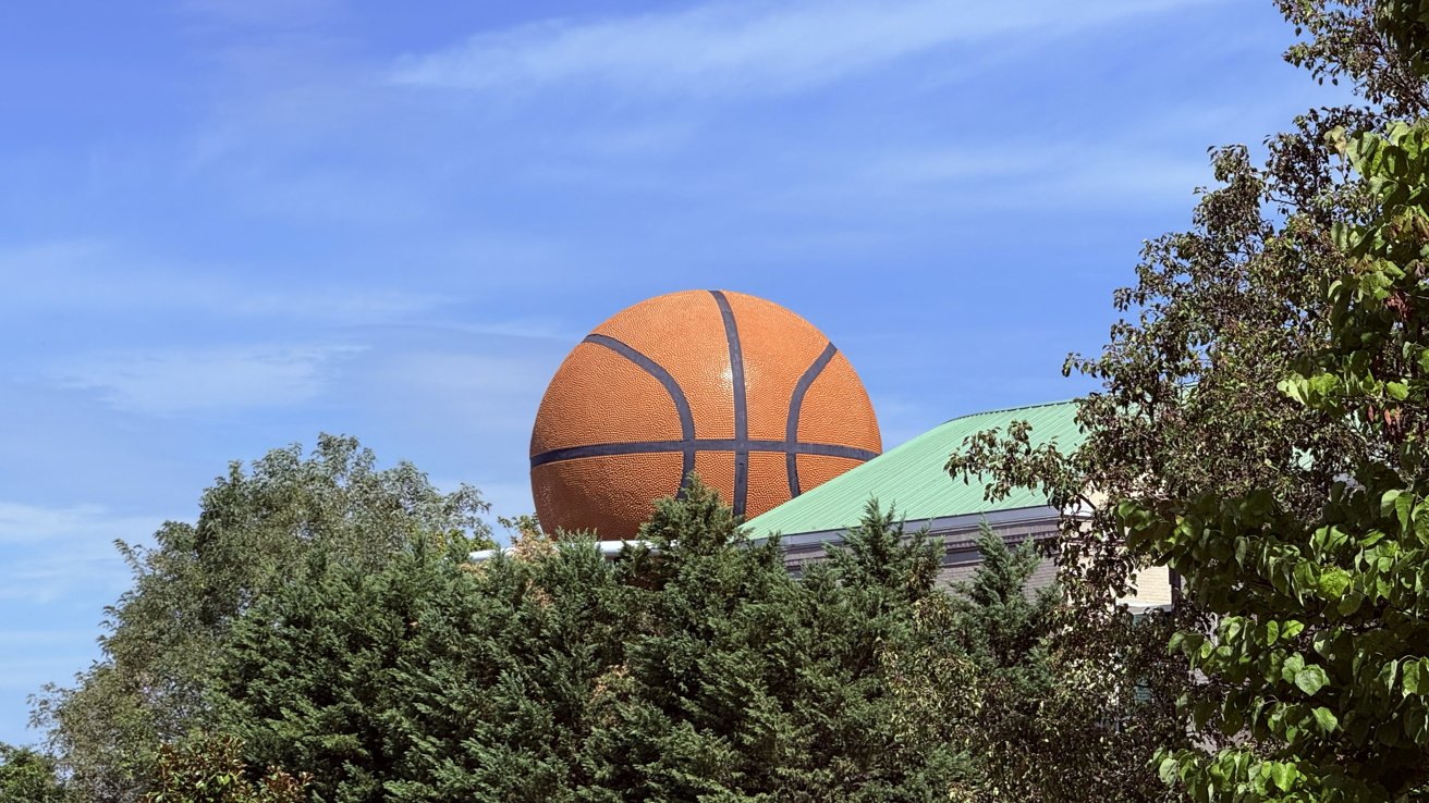 A large basketball sculpture sits atop a building partially obscured by green trees under a clear blue sky.