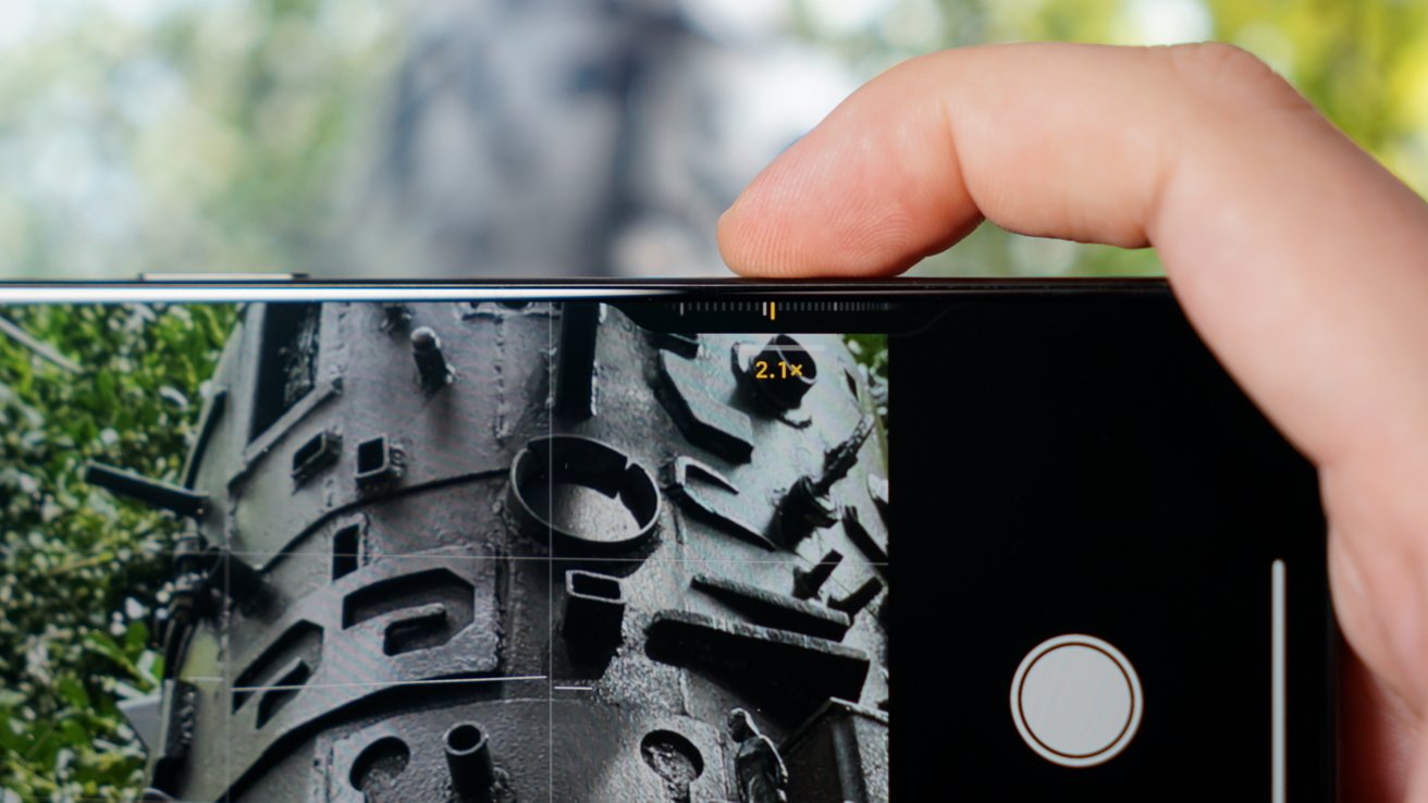 A hand holds a smartphone capturing a photo of a large metal structure with various geometric shapes and protrusions in an outdoor setting.