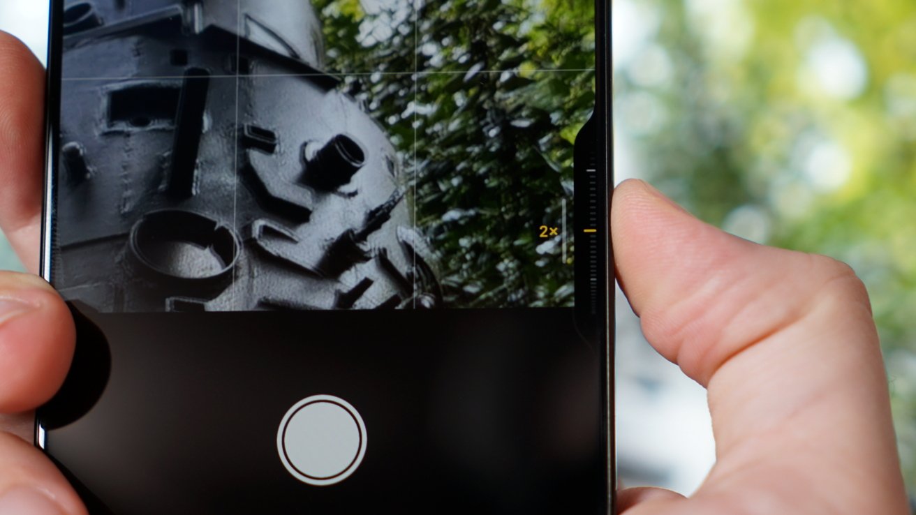 A close-up of a smartphone capturing a photo of a metallic structure outdoors, with greenery in the background.