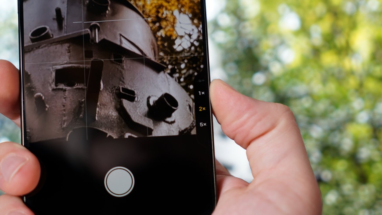 A hand holding a smartphone, capturing a close-up photo of a rusty, industrial structure with green, blurry foliage in the background.