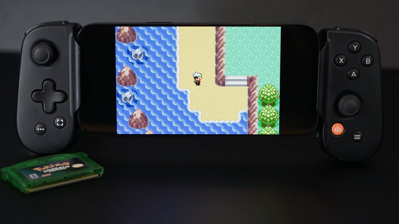 Handheld console displaying pixelated character on a beach with water and rocks. Green game cartridge nearby.