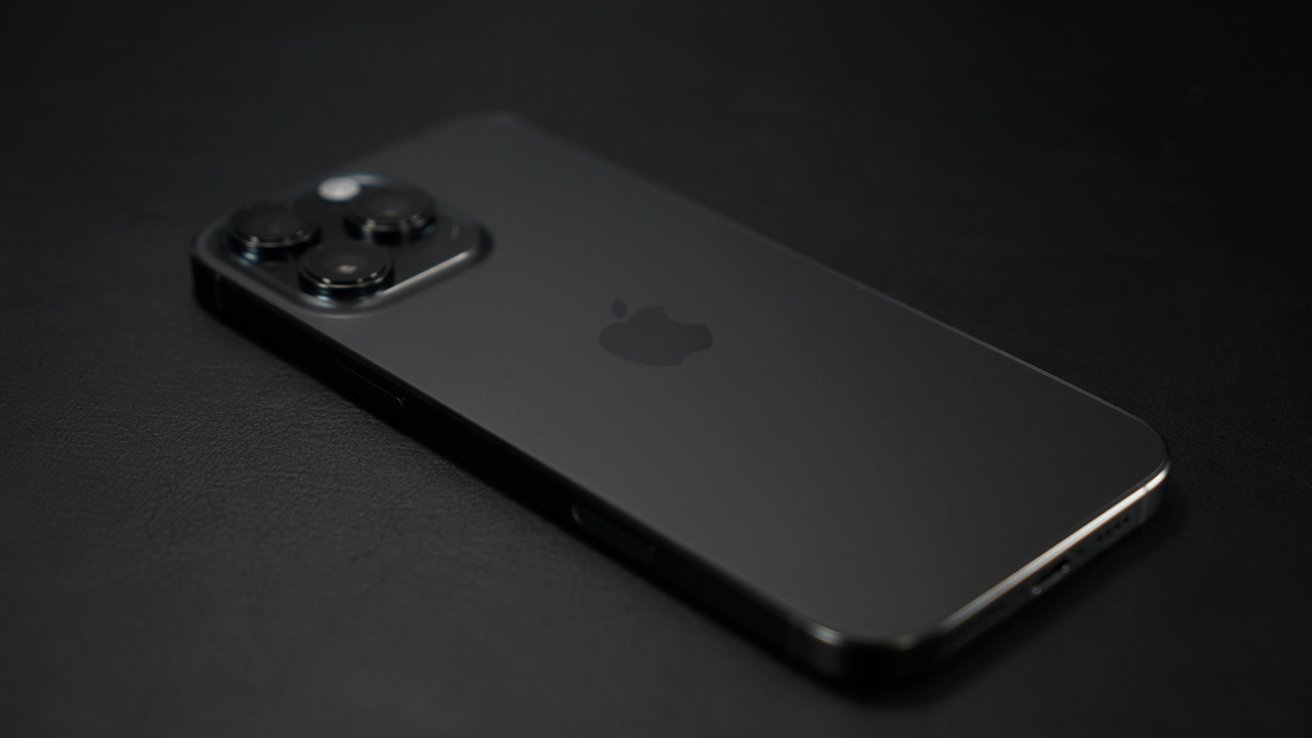 A sleek black smartphone with a rectangular shape and three camera lenses on its back, lying on a dark surface.