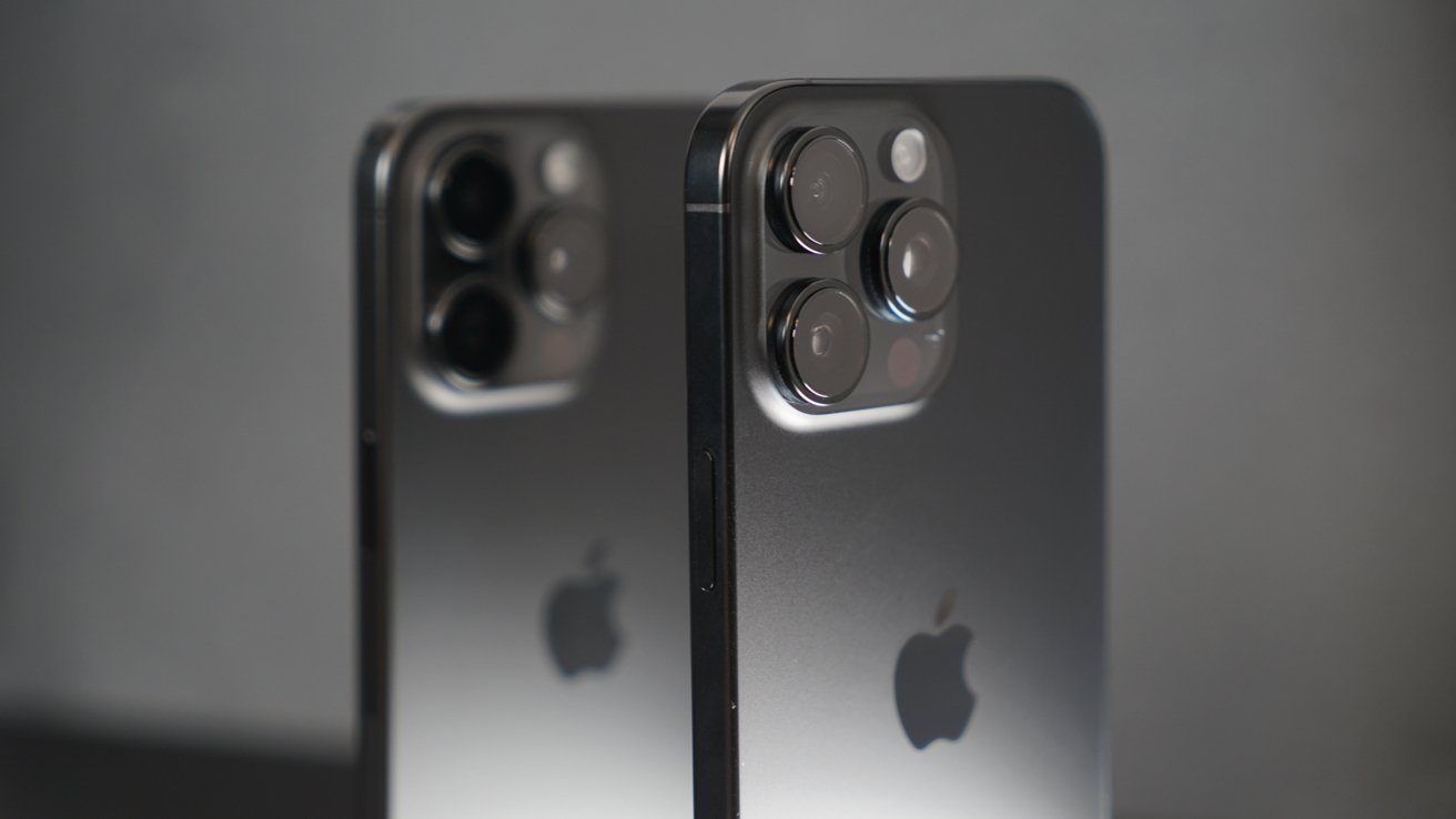 Two dark-colored smartphones with multiple rear cameras, displaying the Apple logo on the back, positioned side by side against a plain background.