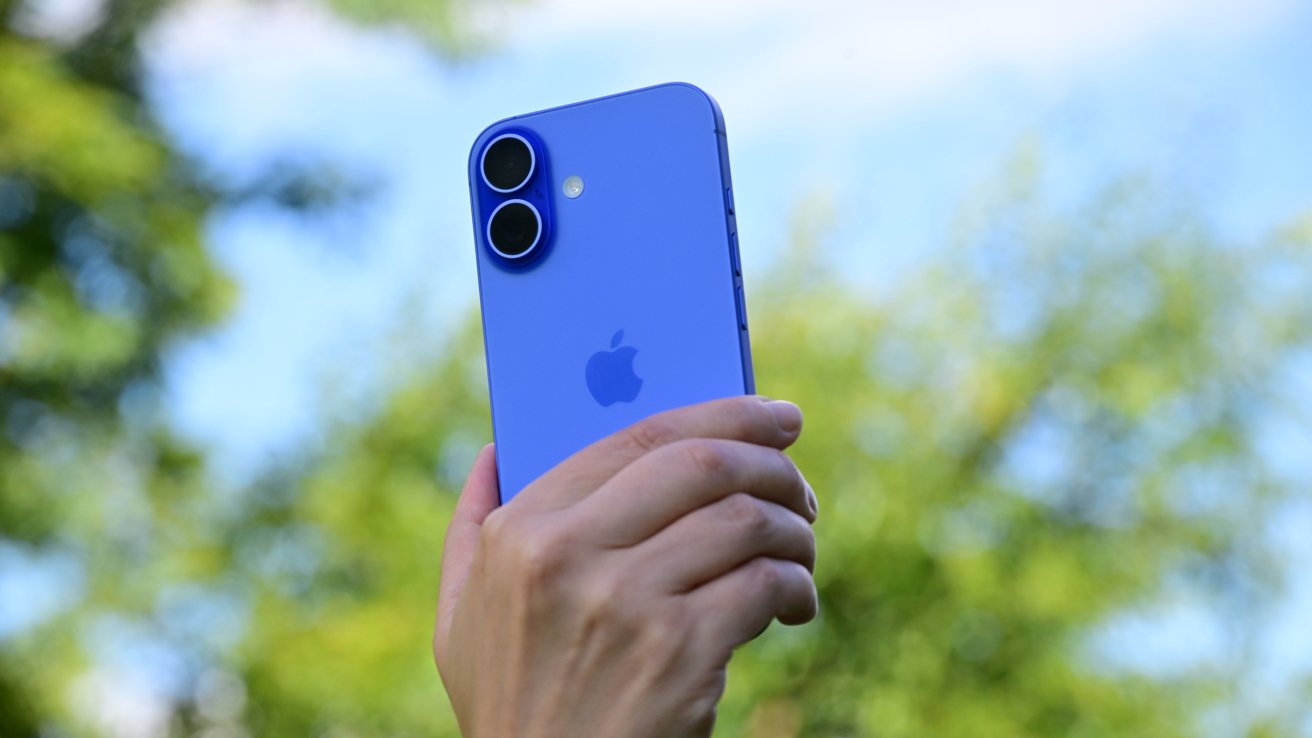 A hand holding a blue smartphone with dual cameras against a blurred background of trees and sky.