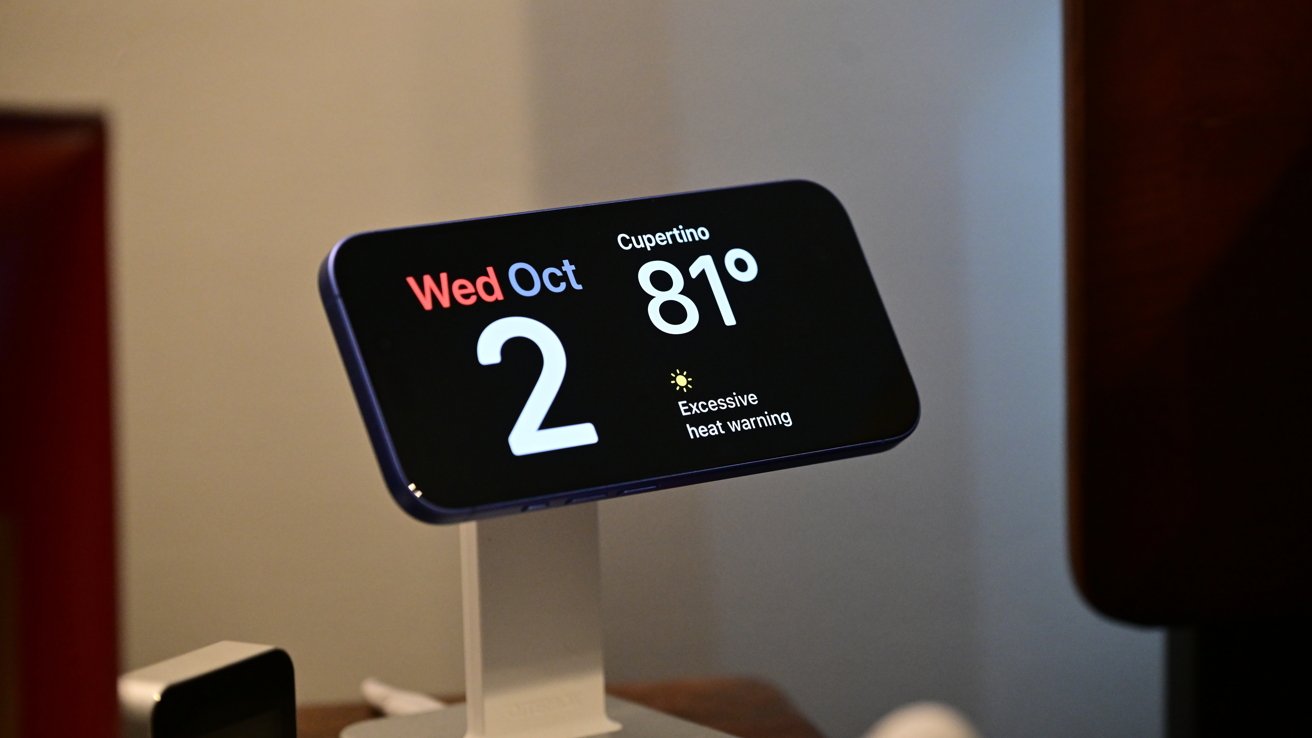 Smartphone displaying date Wednesday, October 2, and Cupertino's temperature at 81F with an excessive heat warning. The phone is mounted on a white stand.