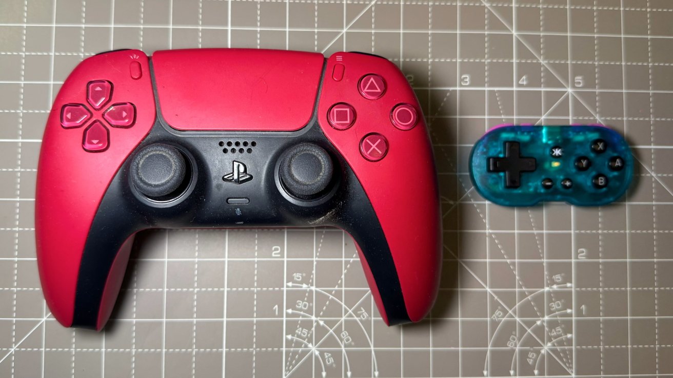 Red and black game controller next to a small, transparent blue controller on a gray cutting mat with measurement lines.