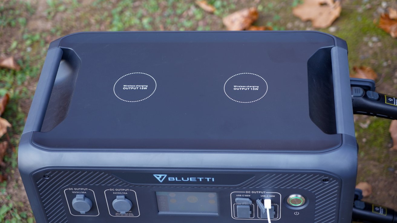 A portable power station with wireless charging pads on top, and various output ports in the front panel, surrounded by fallen leaves on the ground.