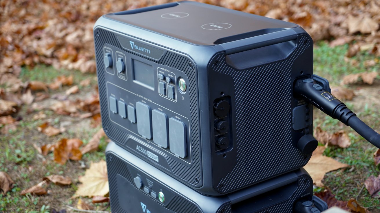 Portable power station with multiple outlets and a connected charging cable, situated outdoors on fallen autumn leaves.