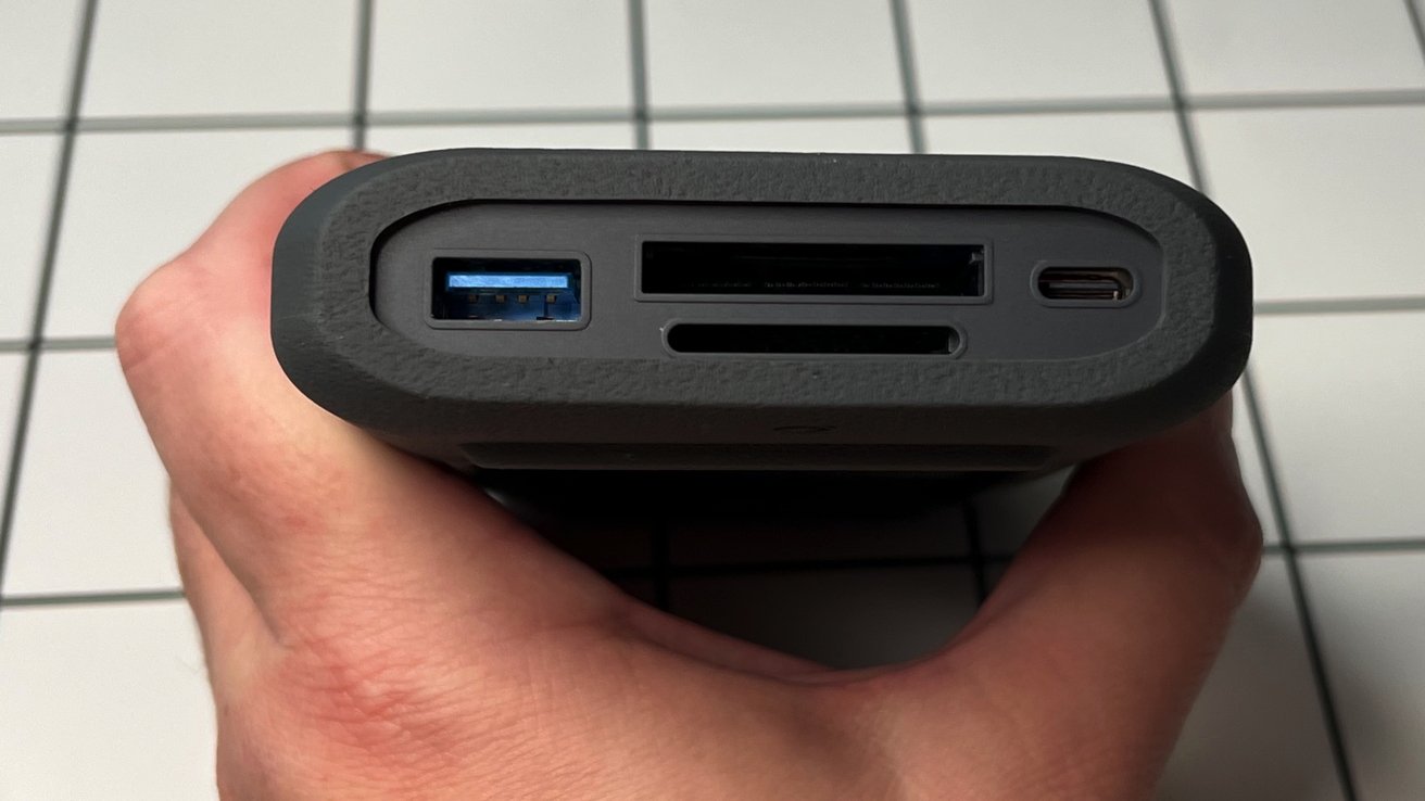 Hand holding a black device with ports including USB, SD card, and USB-C against a tiled background.