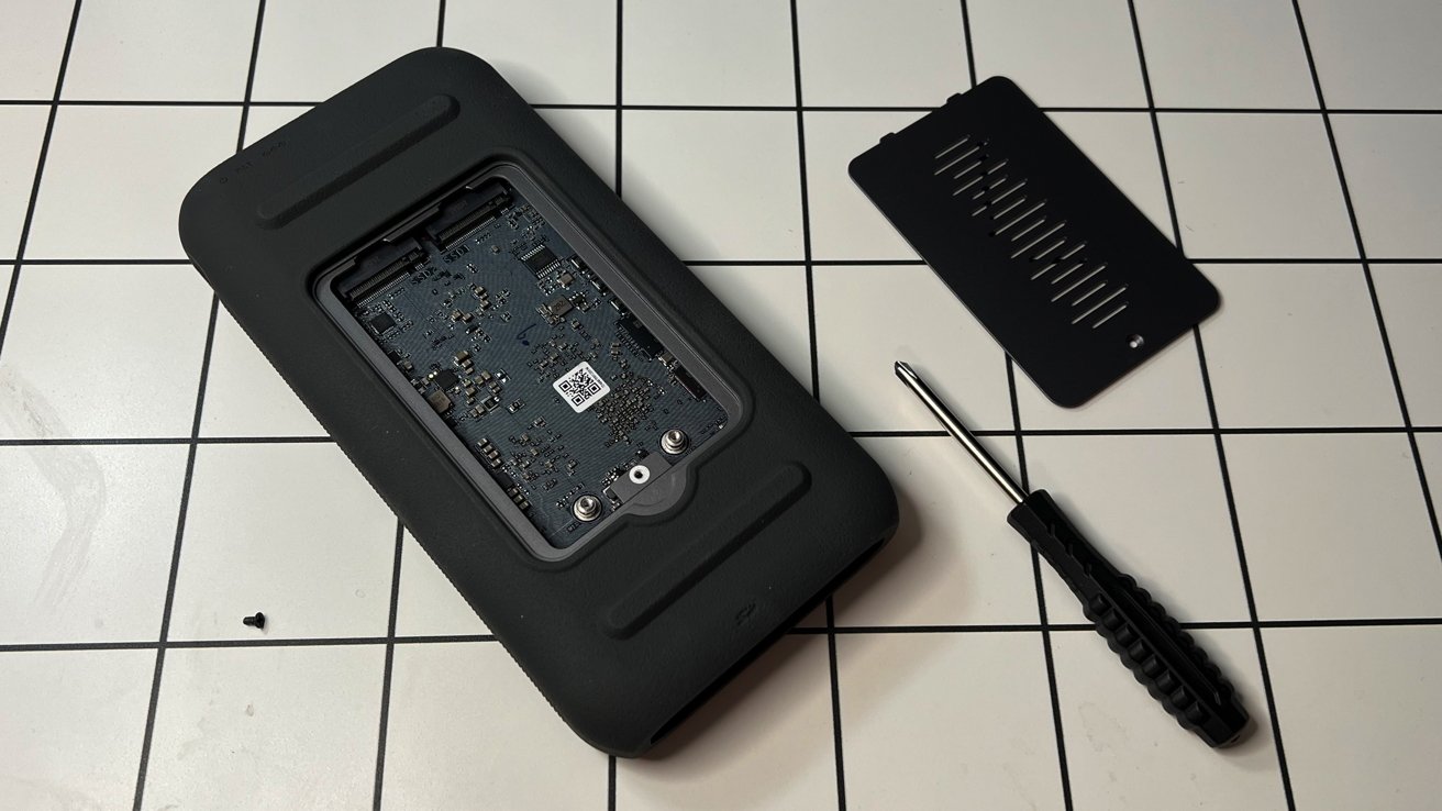 A disassembled black electronic device on a tiled surface, with a circuit board, cover, screwdriver, and small screw nearby.