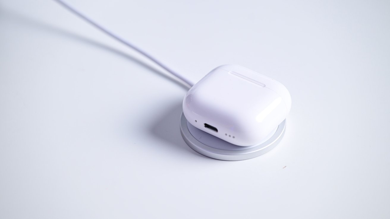 White wireless earbuds case resting on a circular charging pad, with a cable attached, on a light surface.