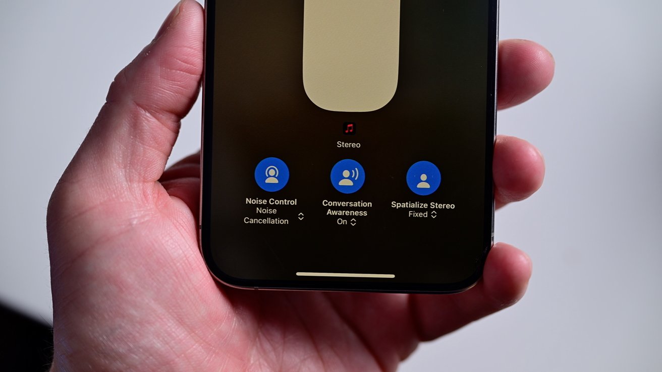 Hand holding a smartphone displaying audio settings: noise control, conversation awareness, and spatialize stereo options on a black background.