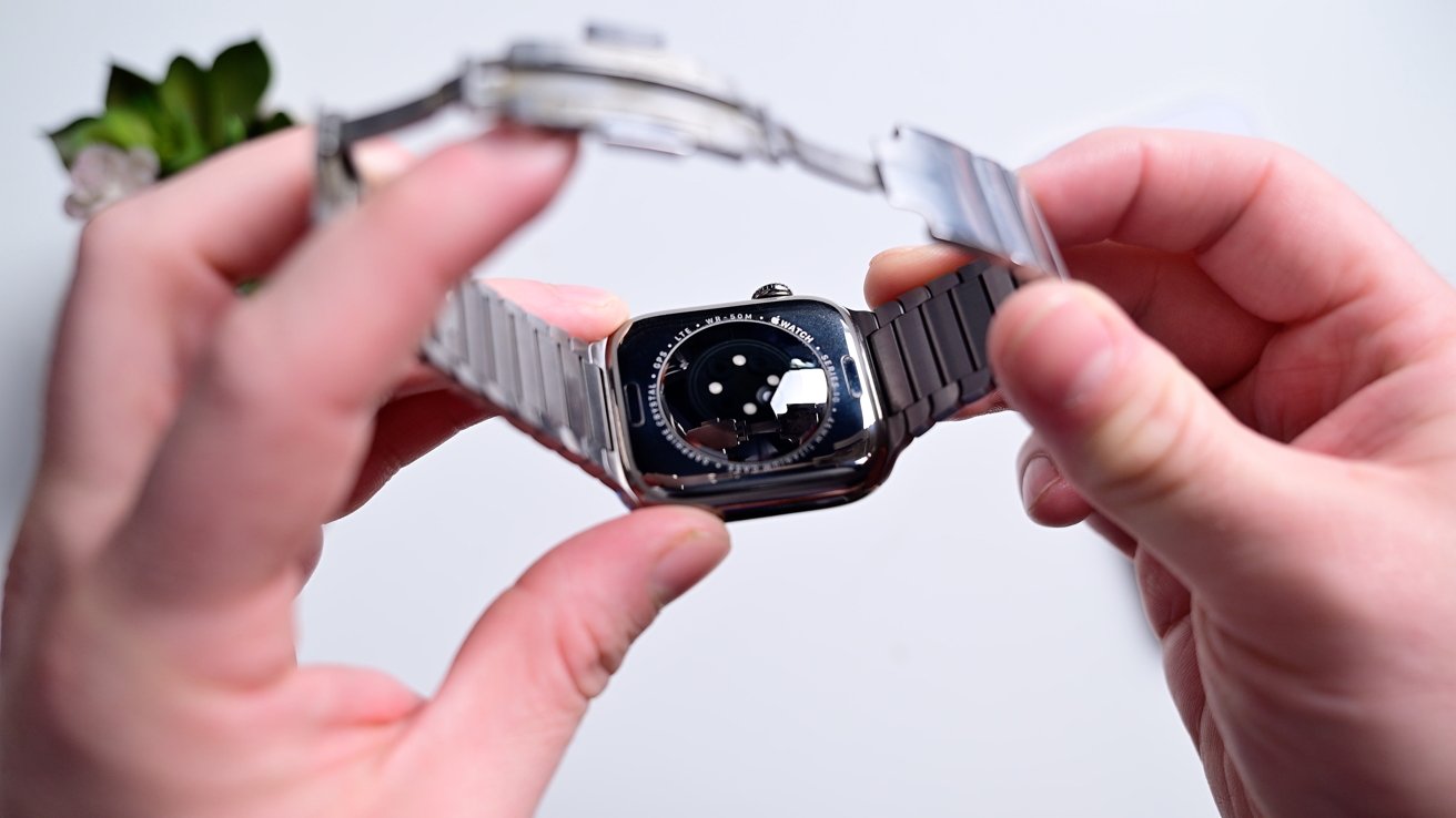 Hands holding a stainless steel smartwatch with a visible sensor on the back, set against a neutral background.