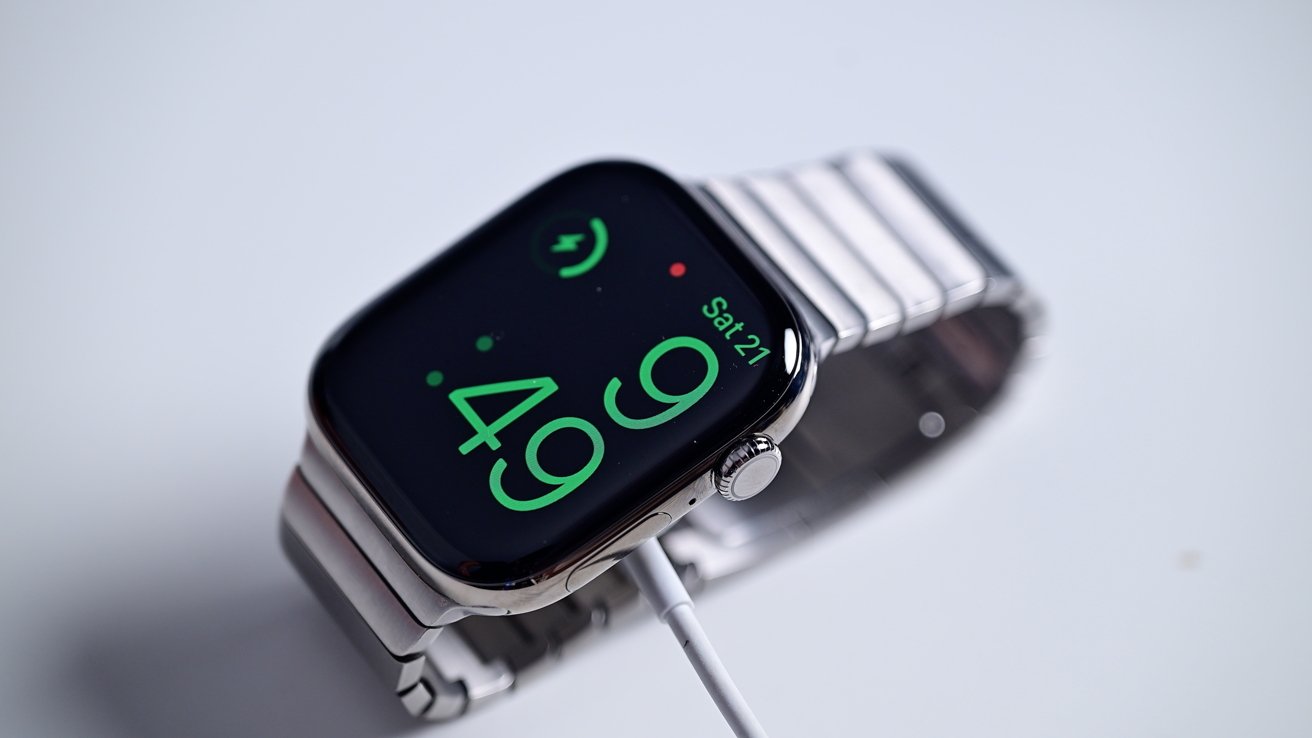 Smartwatch with silver metal band displaying green digital time and date, connected to a charger.