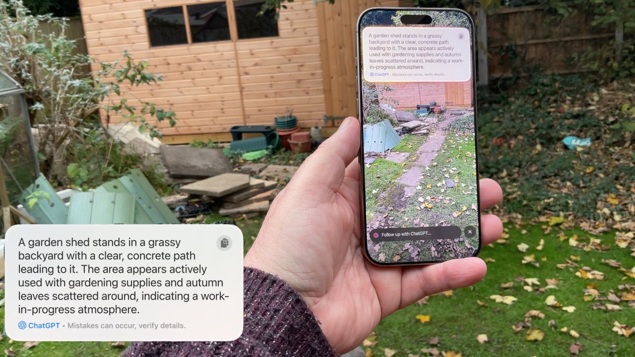 Person holds a phone showing a garden with a shed, path, and scattered leaves, while standing in a backyard with similar features.