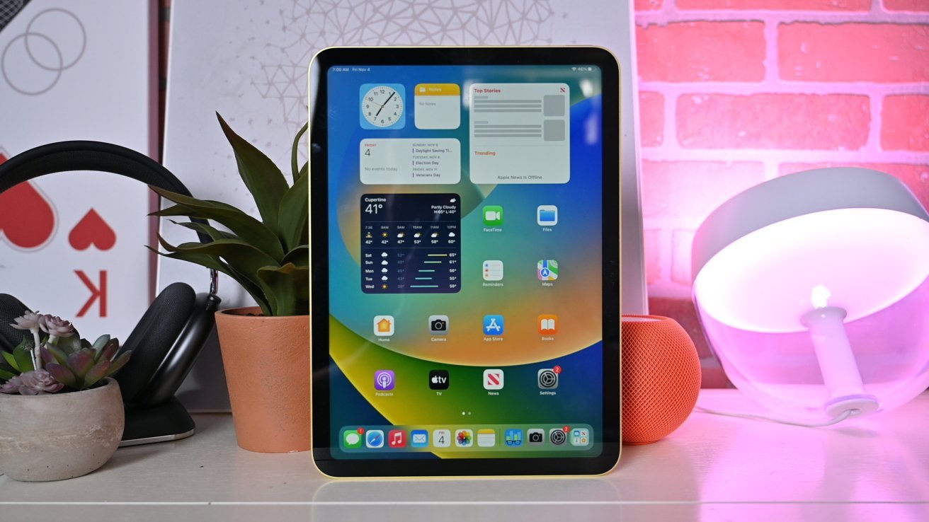 iPad 10th Generation in yellow displaying weather and apps, surrounded by plants, headphones, and a table lamp with pink lighting.