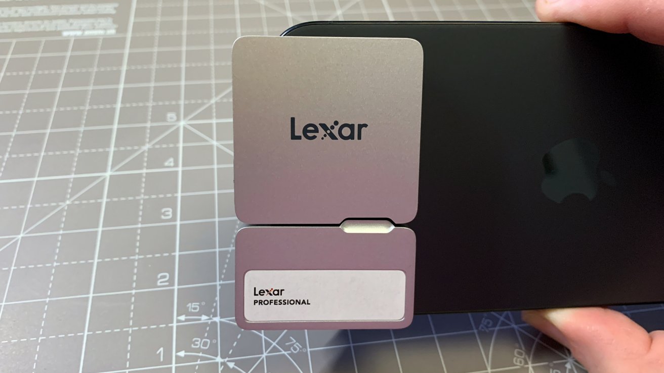 Lexar Professional memory card held by hand in front of a black device with an Apple logo, placed on a cutting mat with measurement markings.