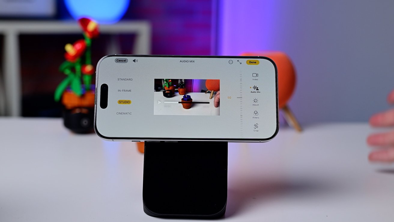 Smartphone on a stand displaying video editing options; background includes a potted plant and orange speaker with purple lighting.