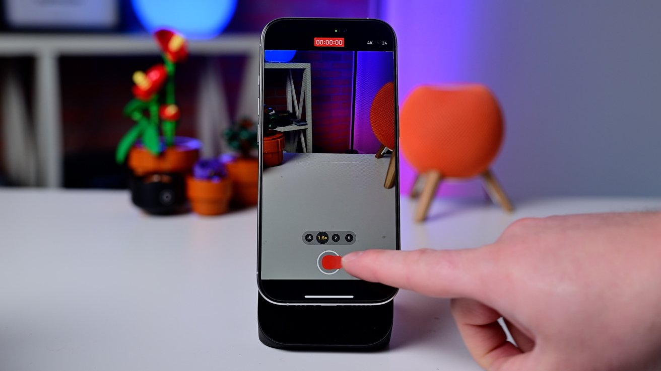 Smartphone in video recording mode on a desk, finger pressing record button, colorful plants and a speaker in the background.