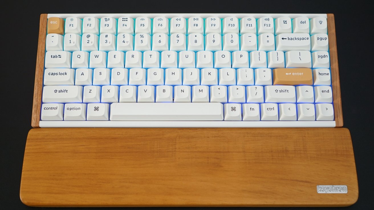 White mechanical keyboard with wooden wrist rest, featuring orange escape and enter keys, placed on a dark surface.