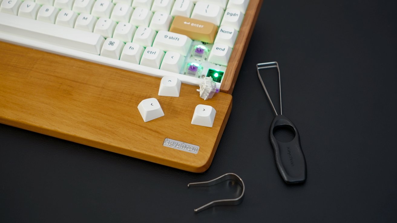 Mechanical keyboard on wooden tray with three removed keycaps, a keycap puller, and a switch puller on black surface.