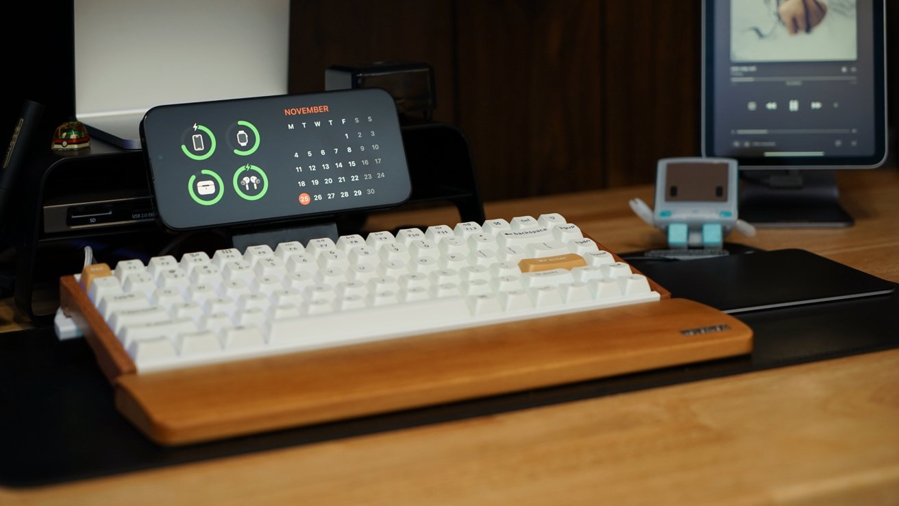 Desk setup with a white keyboard, phone displaying calendar, small robot figure, and tablet playing music, all on a wooden surface.