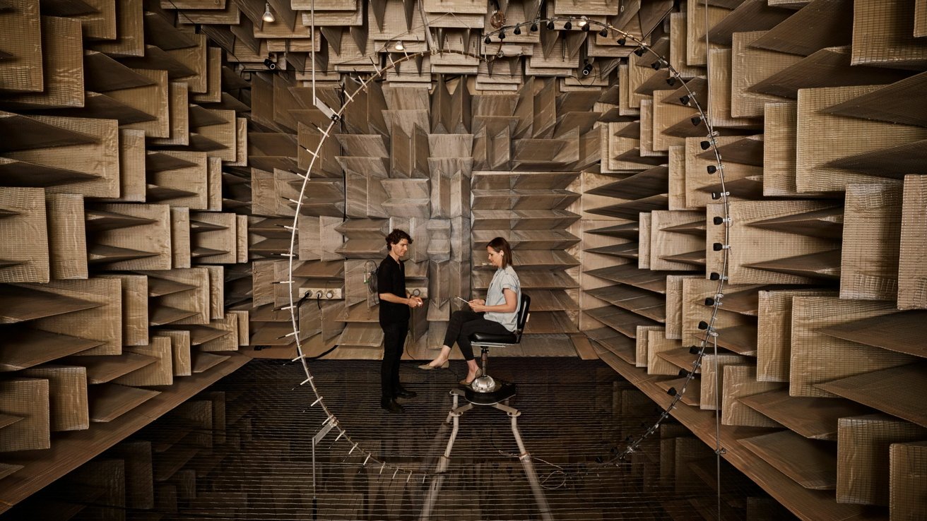 Two people inside an anechoic chamber, surrounded by absorbent panels. One sits on a chair, holding a tablet, while the other stands nearby, amid a circular structure.