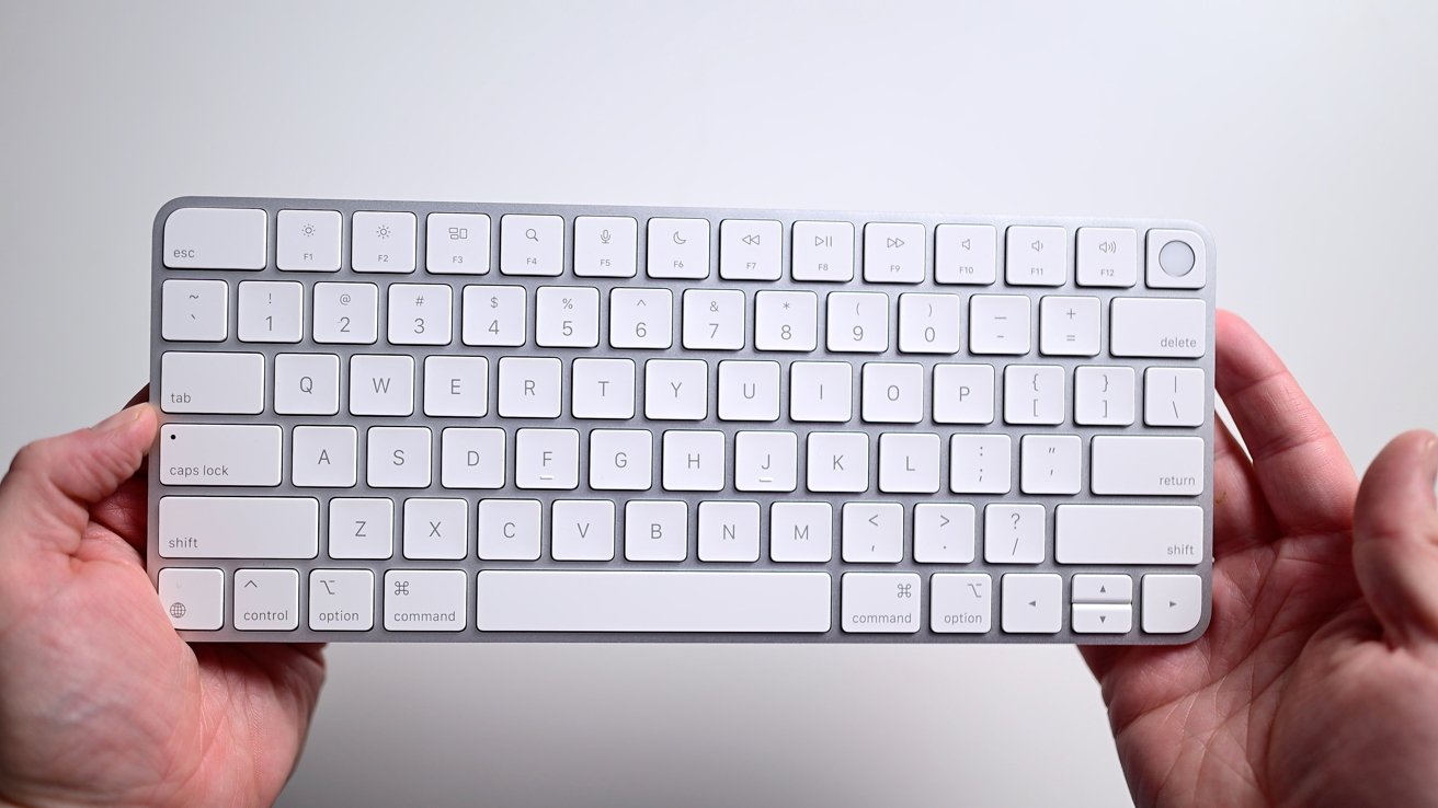 A person holds a wireless white keyboard with black letters and function keys on a gray background.