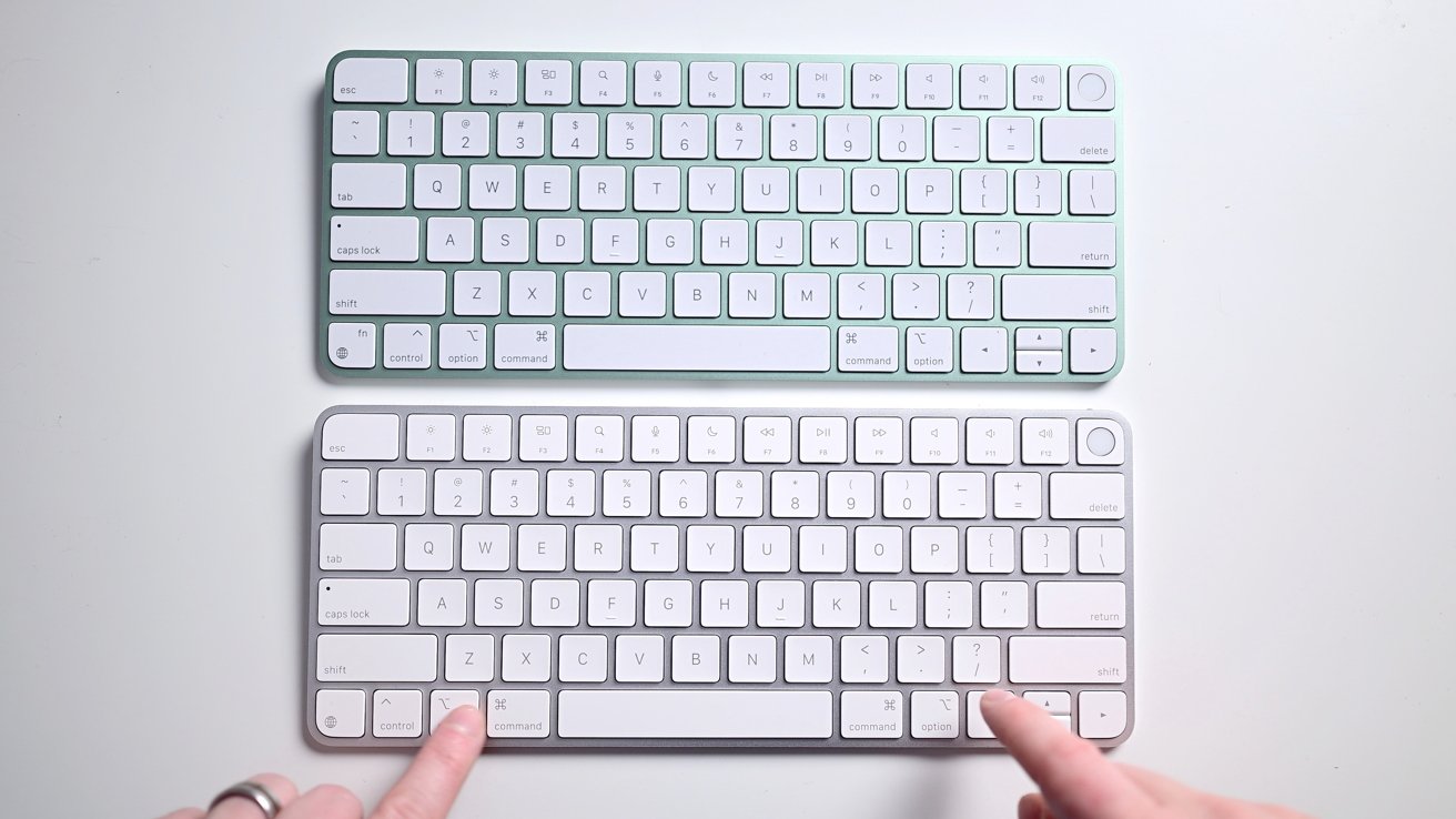 Two compact, white wireless keyboards are placed vertically, with two fingers pointing at keys on the bottom keyboard.